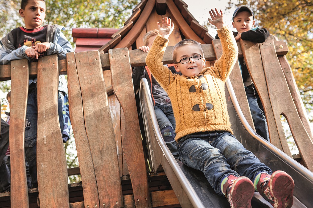 Kinder spielen auf einem Holzklettergerüst inkl. Holzhütte. Zwei Kinder schauen im Hintergrund von oben herunten; ein Junge rutscht im Vordergrund auf der Rutsche herunter.